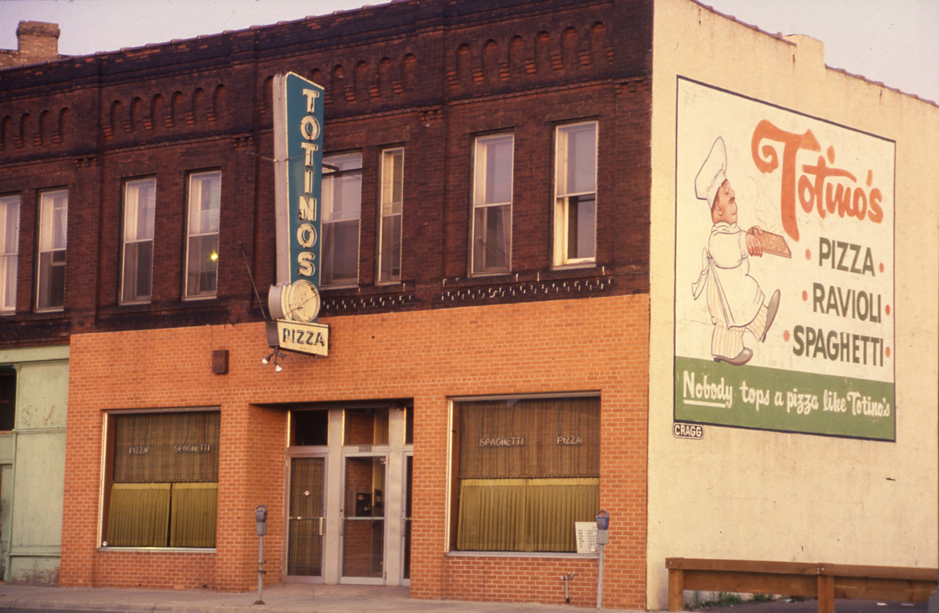 Photo of the exterior of Totino's Pizza shop in 1951. Storefront is orange brick with a vertical neon sign that reads Totino's Pizza. On the right size of the building there is a painted billboard with a Pizza Chef and text that reads Totino's Pizza, Ravioli, Spaghetti, with a slogan at the bottom that reads "Nobody tops a pizza like Totino's".